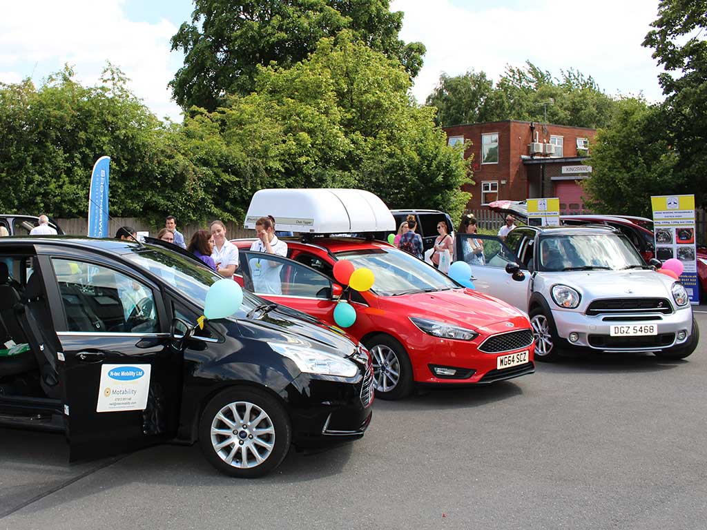 A Driving Mobility open day with three cars and people standing next to them.